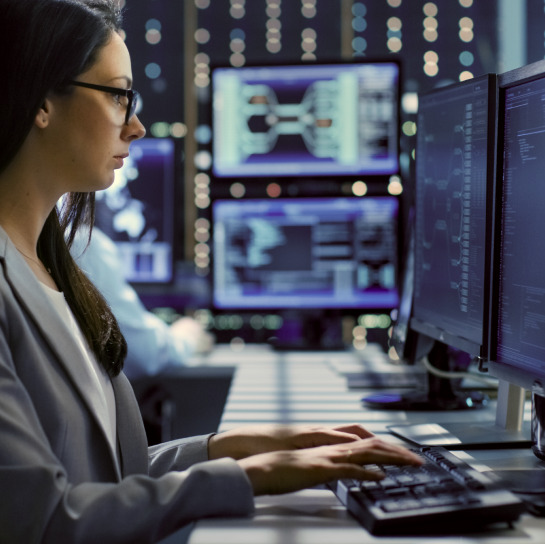 Female cybersecurity analyst reviewing data through a dual monitor screen configuration in an office at night