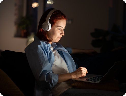 A woman wearing headphones, focused on her laptop, illuminated by soft light in a nighttime setting waiting for artificial intelligence results