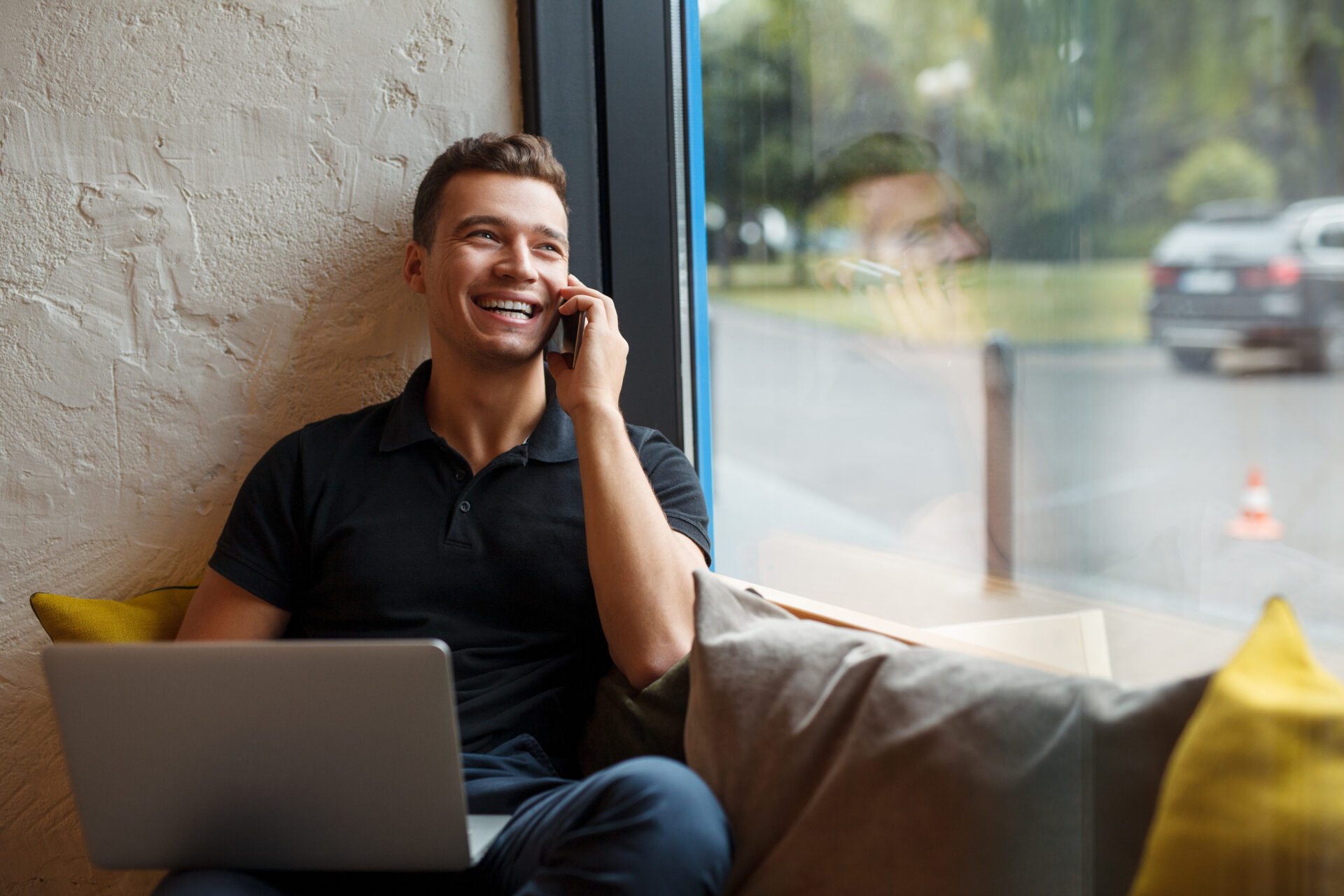 Smiling male using his laptop and connecting with a customer through the phone at a cafe