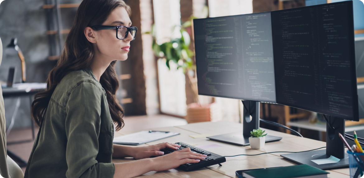 Female worker with glasses reviewing code in a dual screen monitor in an office like setting