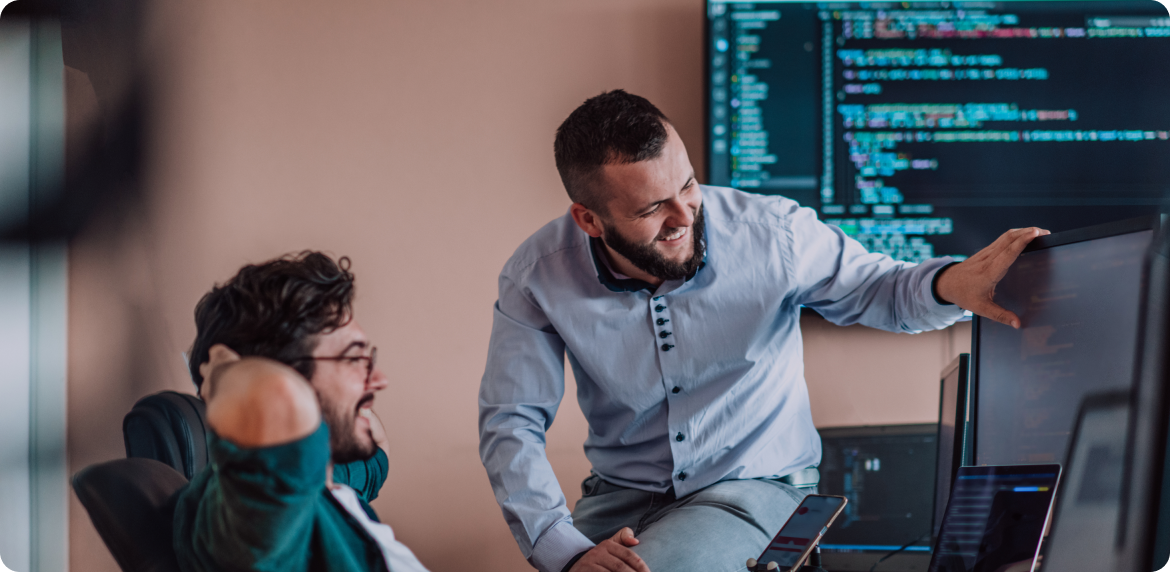 Two male coworkers reviewing code in a laptop and a monitor screen in an office