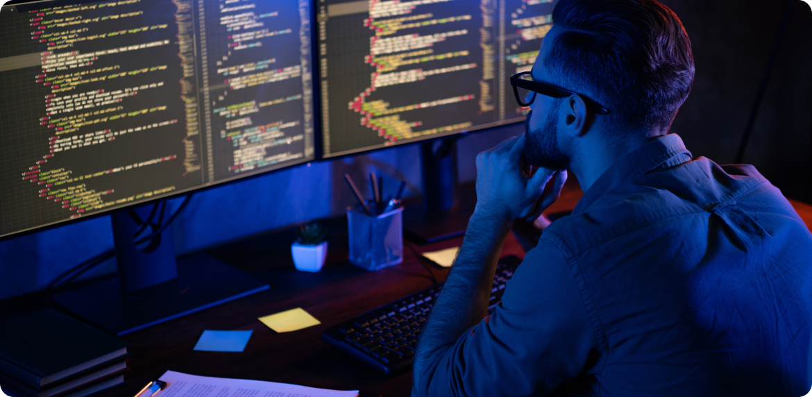 Male developer with glasses reviewing code in a dual screen monitor at night