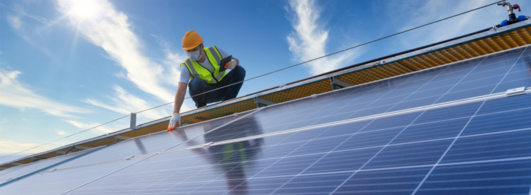 A man in a hard hat and safety vest installs a solar panel under clear blue skies.