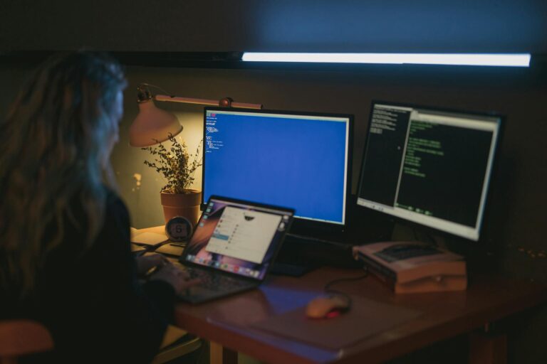 A woman working at a desk with two computer screens, focused on her tasks and surrounded by a professional environment.