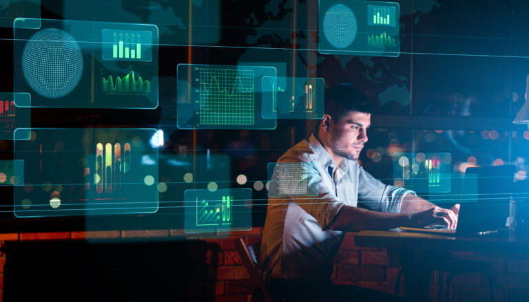 An artificial intelligence expert at a table using a laptop, with a monitor beside him showcasing graphs and analytical information.